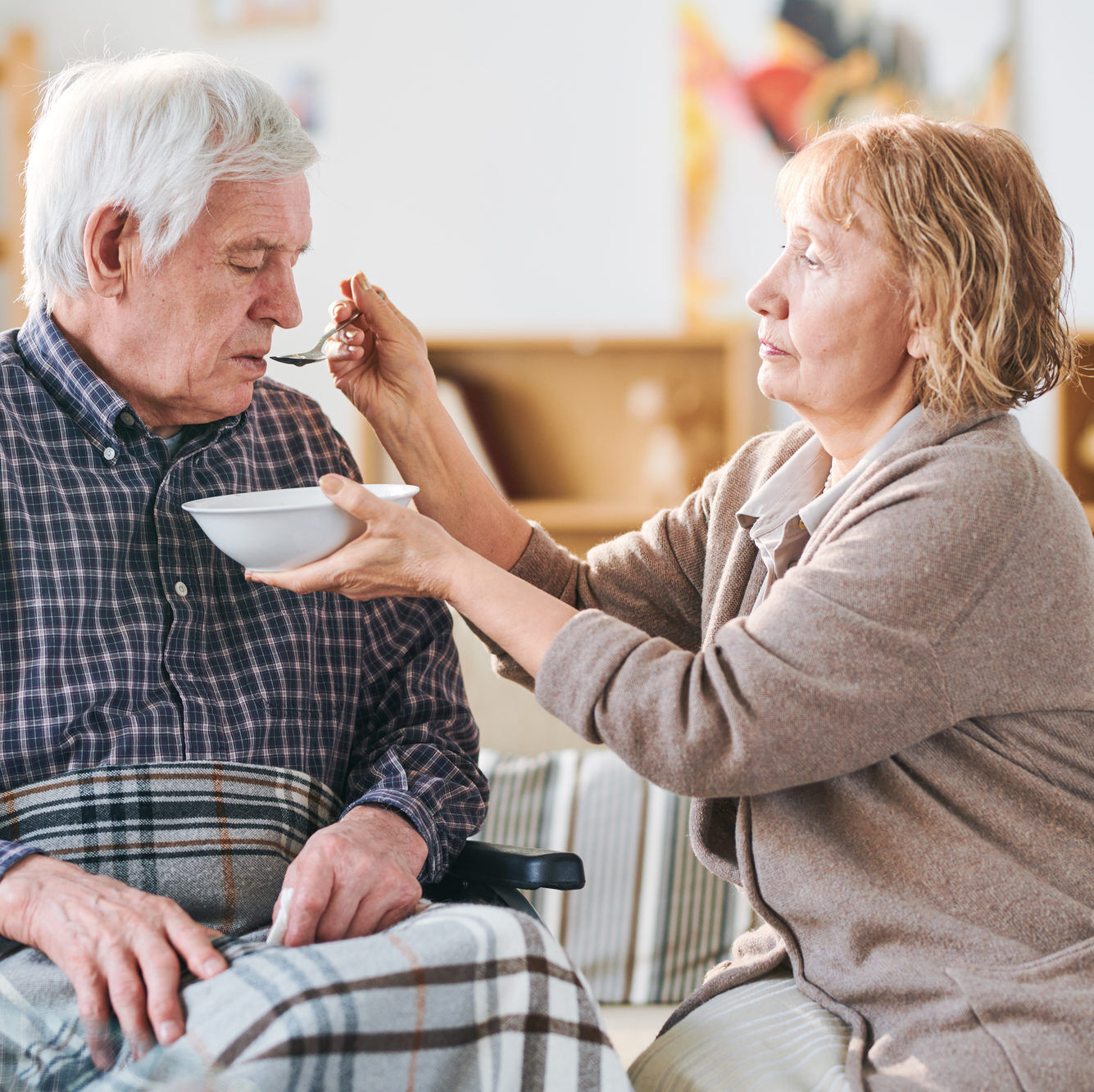 Aged casual woman holding bowl with soup and spoon by her disable husband mouth while helping him to eat dinner