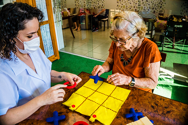 Senhora idosa, sentada à mesa junto com profissional especializada, jogando jogo da velha feito em tecido, durante atividade de terapia ocupacional