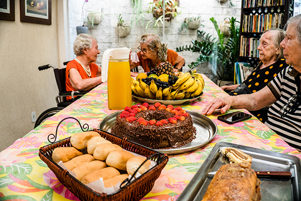 Mesa com pães, bolo, suco e flores. Ao fundo, duas senhoras idosas moradoras do SoLAr de Itaipú colocam o papo em dia.