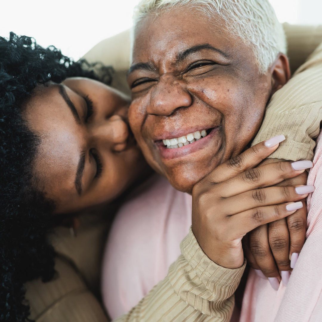 Uma jovem negra abraça sua avó com carinho, pelas costas, dando um beijo na sua bochecha. A avó, uma senhora negra sorridente na faixa dos 70 anos, veste uma blusa de tricot rosa. A imagem transborda amor, união e a forte conexão entre avó e neta. Relação de intergeracionalidade.