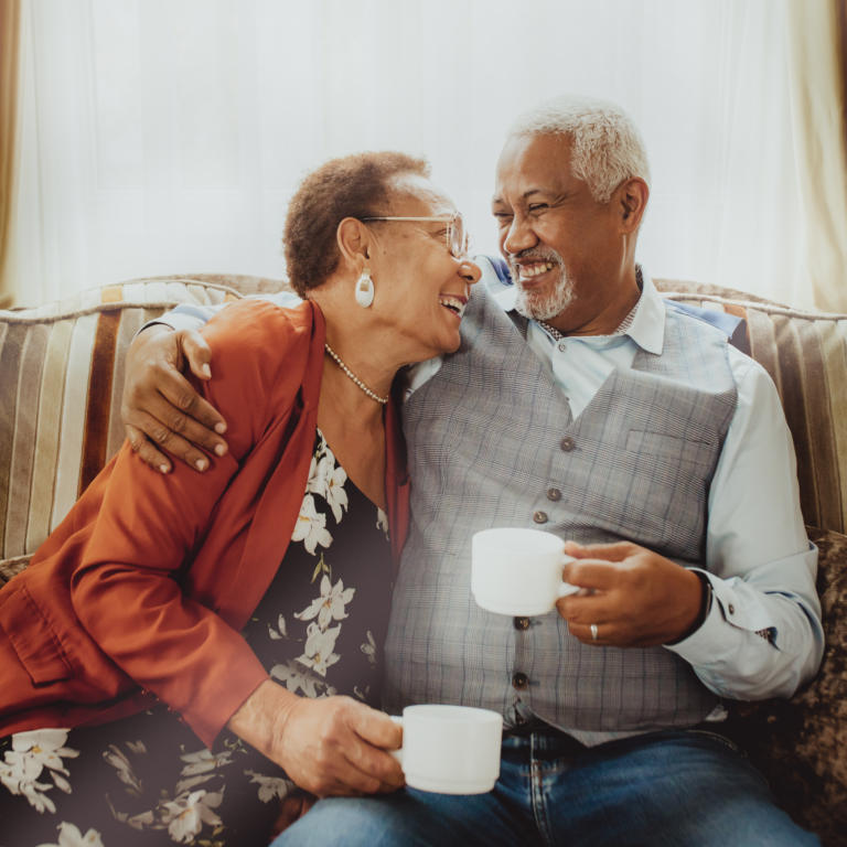 Casal de idosos de pele negra, sorrindo e se entreolhando de forma afetuosa enquanto bebem café no sofá da sala de estar, simbolizando a importância de momentos de convivência e saúde na terceira idade.