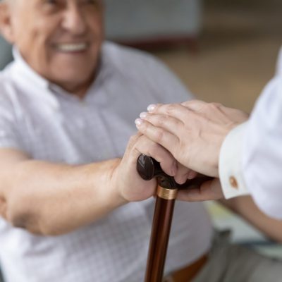 Close up of nurse caregiver palms covering hand of smiling senior patient holding cane. Sick old man struggle with trauma consequence make success on rehabilitation therapy with help support of doctor