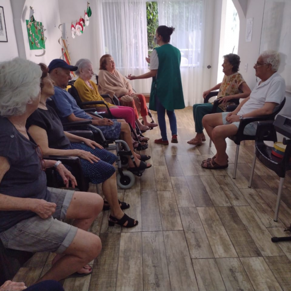 Sete idosos sentados em semi-círculo participando de atividade de música com profissional que está em pé no centro