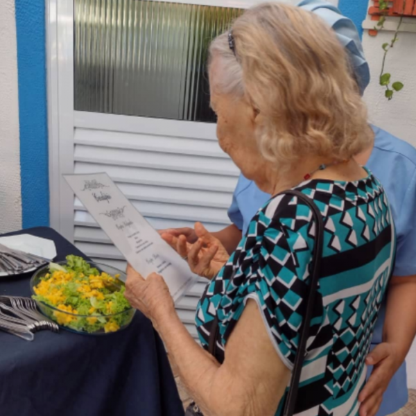 Senhora idosa residente do Longévité lendo cardápio para escolher o que deseja, com o apoio de uma profissional do residencial
