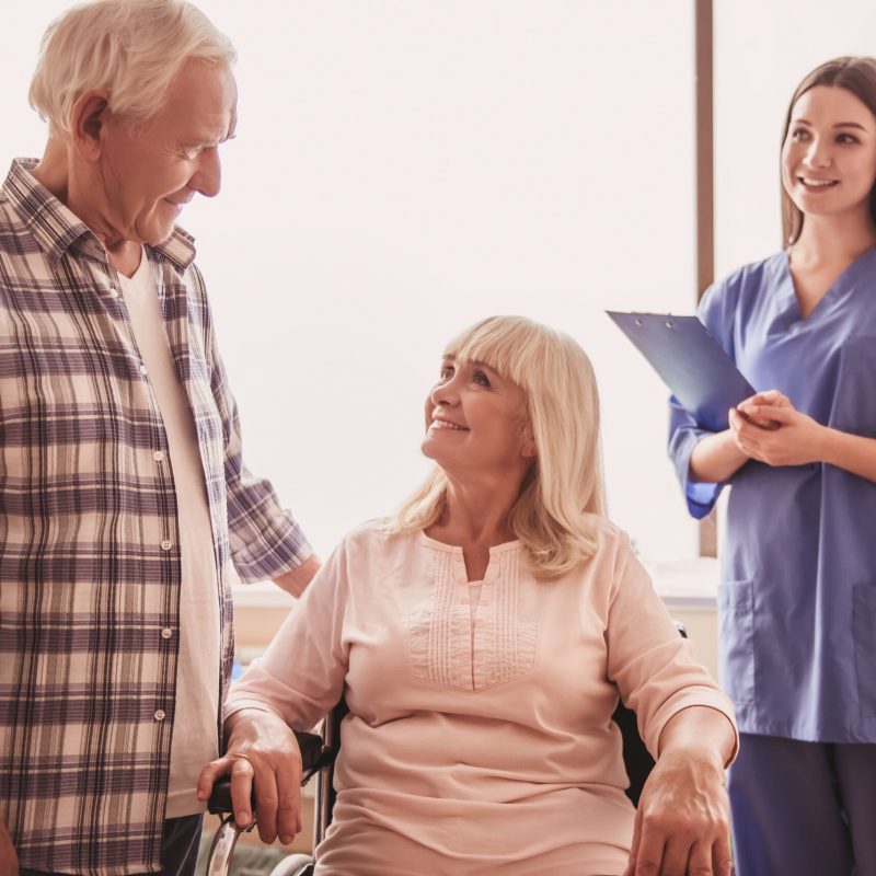 Beautiful old lady is talking to her husband while sitting in the wheelchair in hospital ward, nurse is standing near, all are smiling