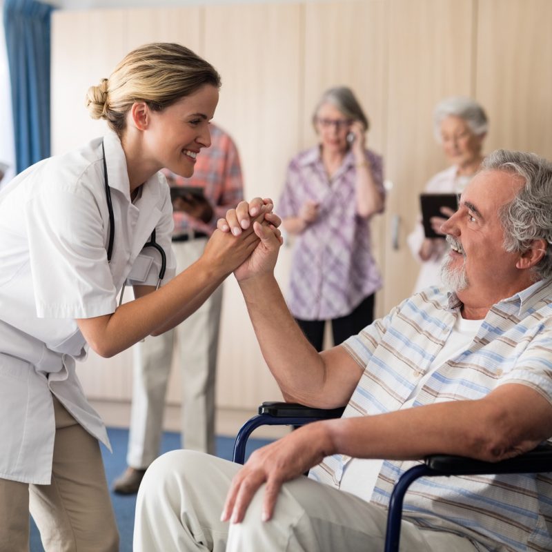 Smiling female doctor holding hands with disabled senior man sitting on wheelchair at retirement home